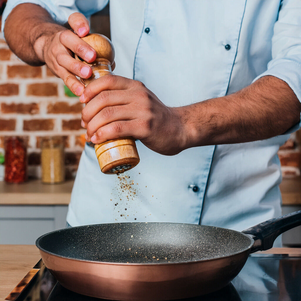 Hænder kværner frisk peber med en trækværn over en non-stick stegepande på komfuret; kokkens hvide jakke og murstensvæg er synlige i baggrunden.