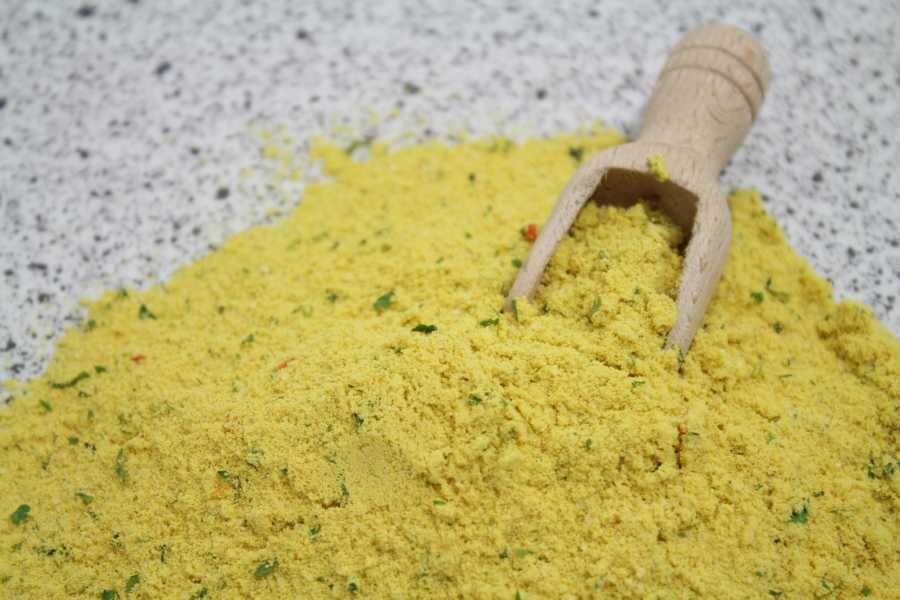 Nærbillede af gul krydderiblanding i pulverform med små grønne urtestykker og en træske på bordet