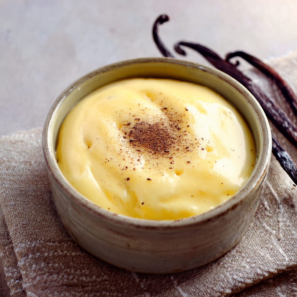 Nærbillede af fløjlsblød vaniljecreme i en keramisk skål, pyntet med et let drys kanel; vaniljestænger ligger ved siden af.