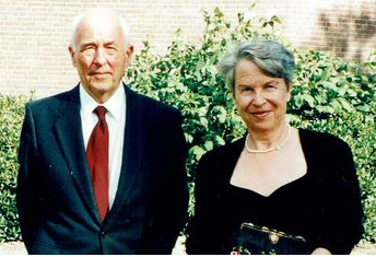 To voksne personer iført formelt tøj: en mand i jakkesæt med rødt slips og en kvinde i sort kjole med taske står udendørs foran grønne buske.