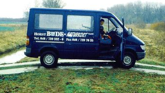 Blå varevogn parkeret på en ujævn markvej med firmalogo og telefonnummer på siden; fører synlig gennem førersiden.