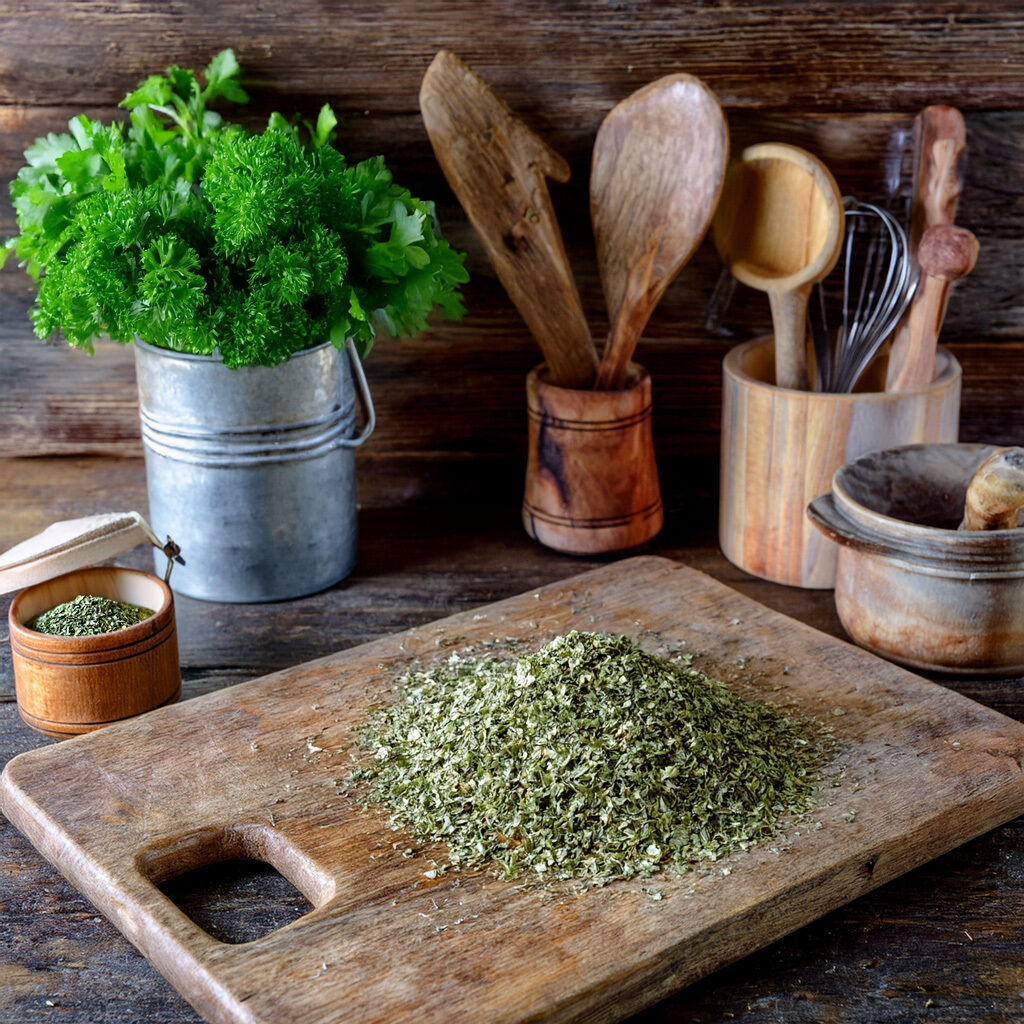 A wooden kitchen counter with a pile of dried parsley, fresh parsley in a metal pot, and rustic wooden utensils