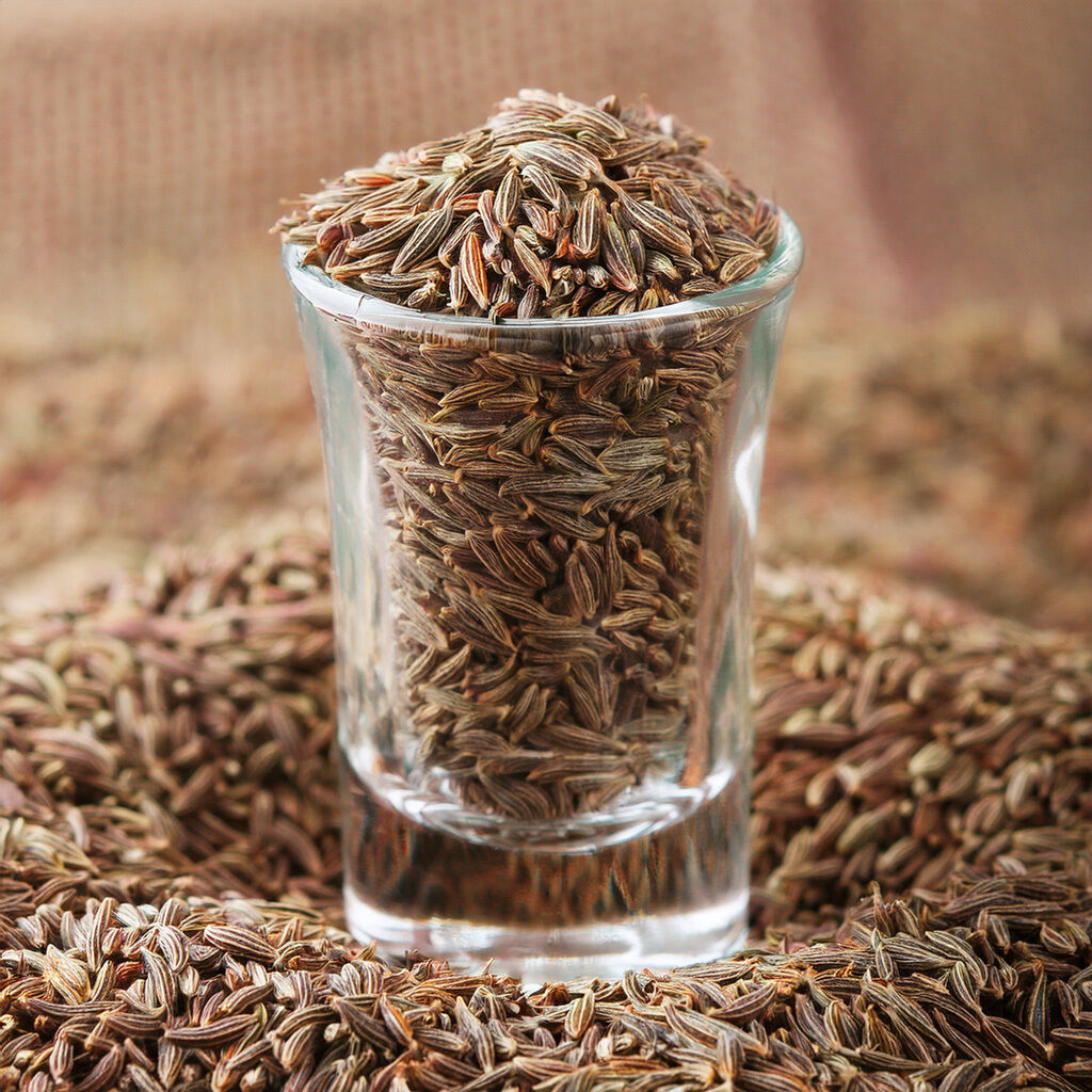Nærbillede af et lille glas fyldt med hele kommenfrø (karvefrø) med spredte frø omkring, brune lange krydderifrø i fokus.