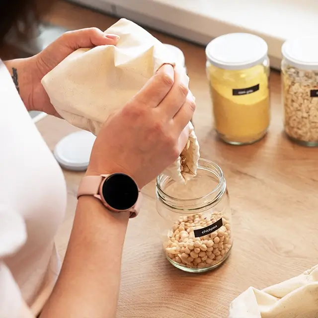 Hænder hælder kikærter fra en stofpose i en glasbeholder på et køkkenbord, med andre glasbeholdere med krydderier i baggrunden.
