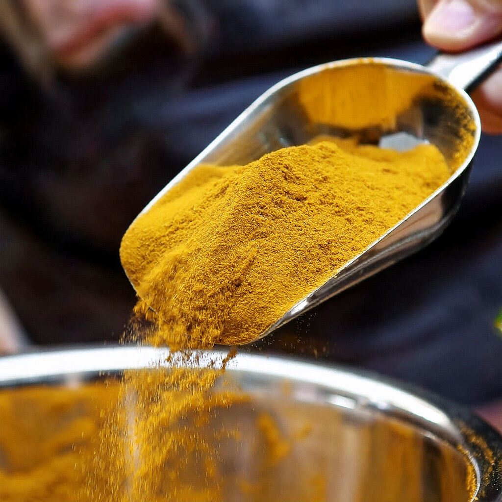 Metalskovl hælder gult krydderipulver (f.eks. gurkemeje) ned i en skål