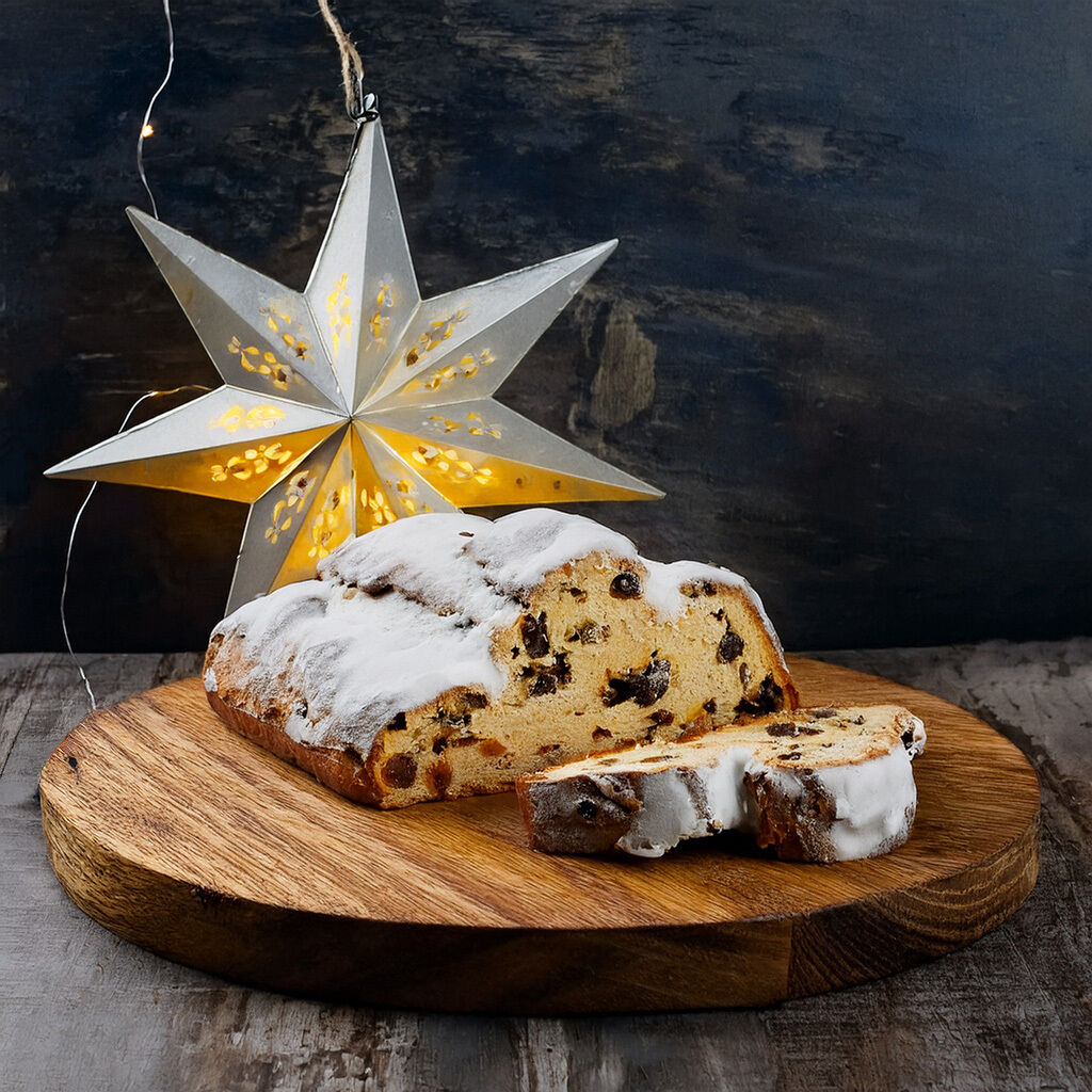 Ein festlicher Laib Weihnachtsstollen, bestreut mit Puderzucker, serviert auf einem Holzbrett neben einer leuchtenden Sternlaterne.