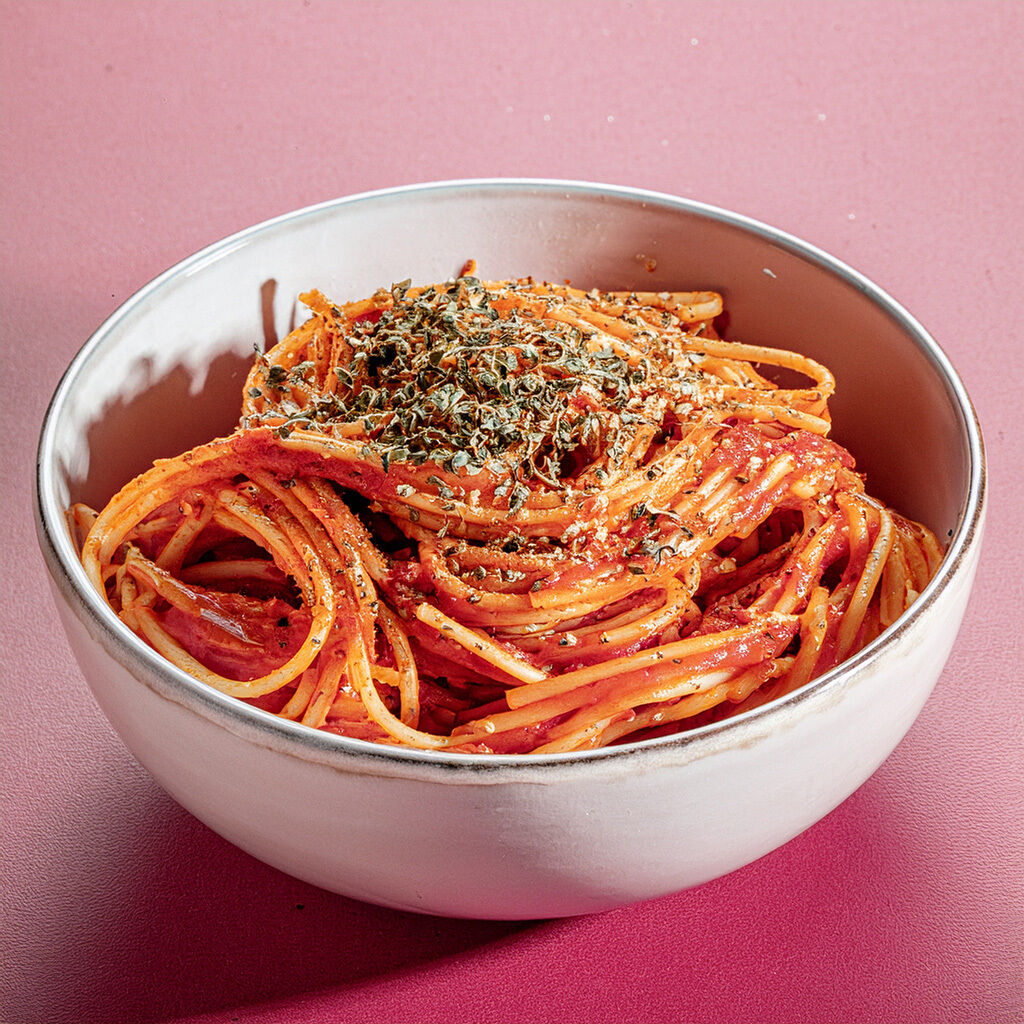 Hvid skål med spaghetti i rød tomatsauce, drysset med tørrede krydderurter