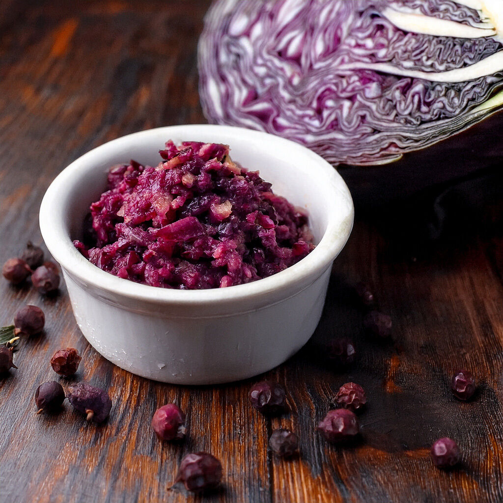 A white bowl filled with seasoned red cabbage beside a halved red cabbage and scattered juniper berries on a wooden surface.
