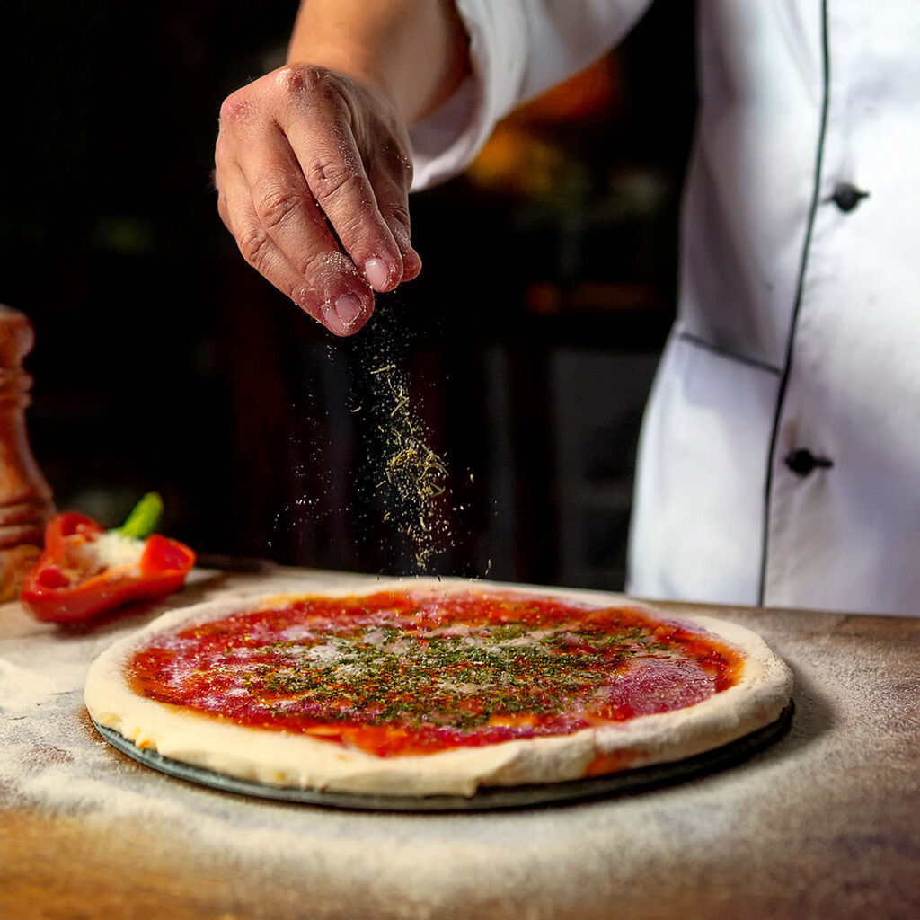 Kok drysser urter på en frisk tomatpizzadej på et træbord.