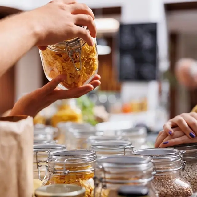 Nærbillede af hænder, der holder et patentglas fyldt med cornflakes over en række lukkede glasbeholdere i en selvbetjeningsbutik