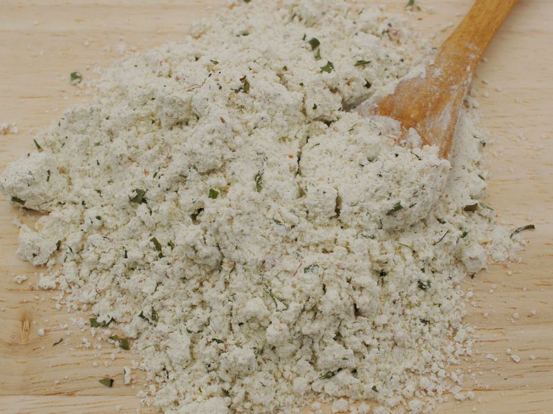 Nærbillede af hvidt krydret paneringspulver med grønne urteflager og en træske på et lyst træbord.