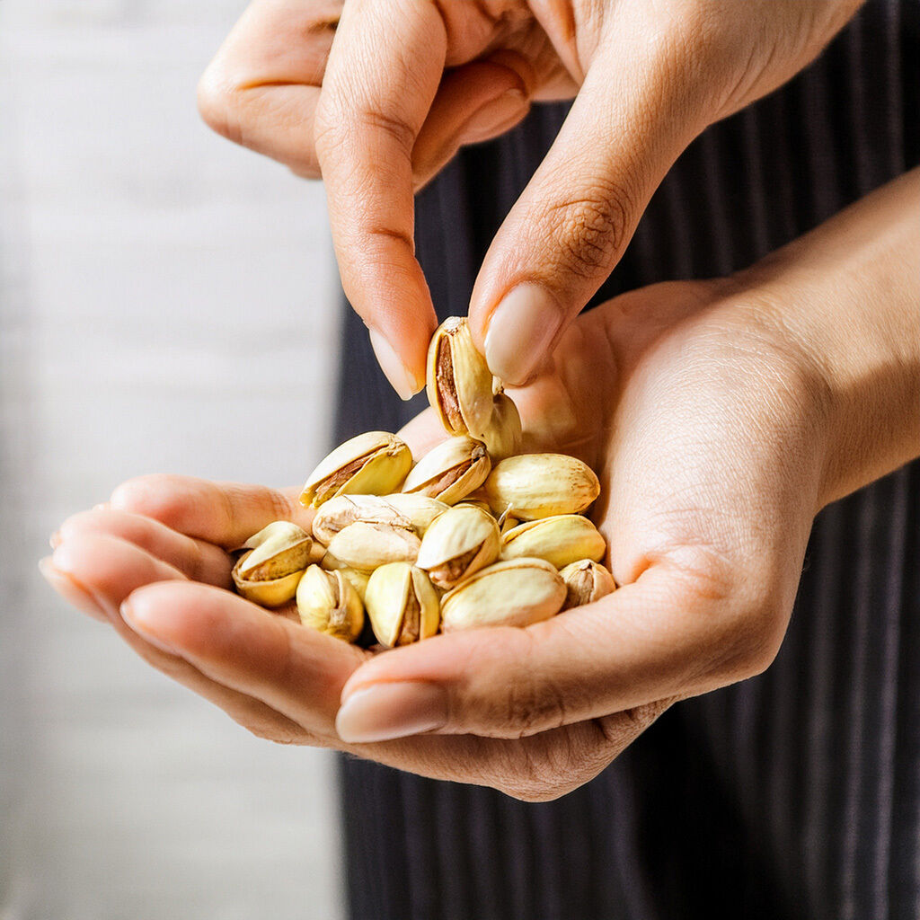 Nahaufnahme: Hände halten eine Portion Pistazien, eine Hand nimmt eine geöffnete Pistazie heraus