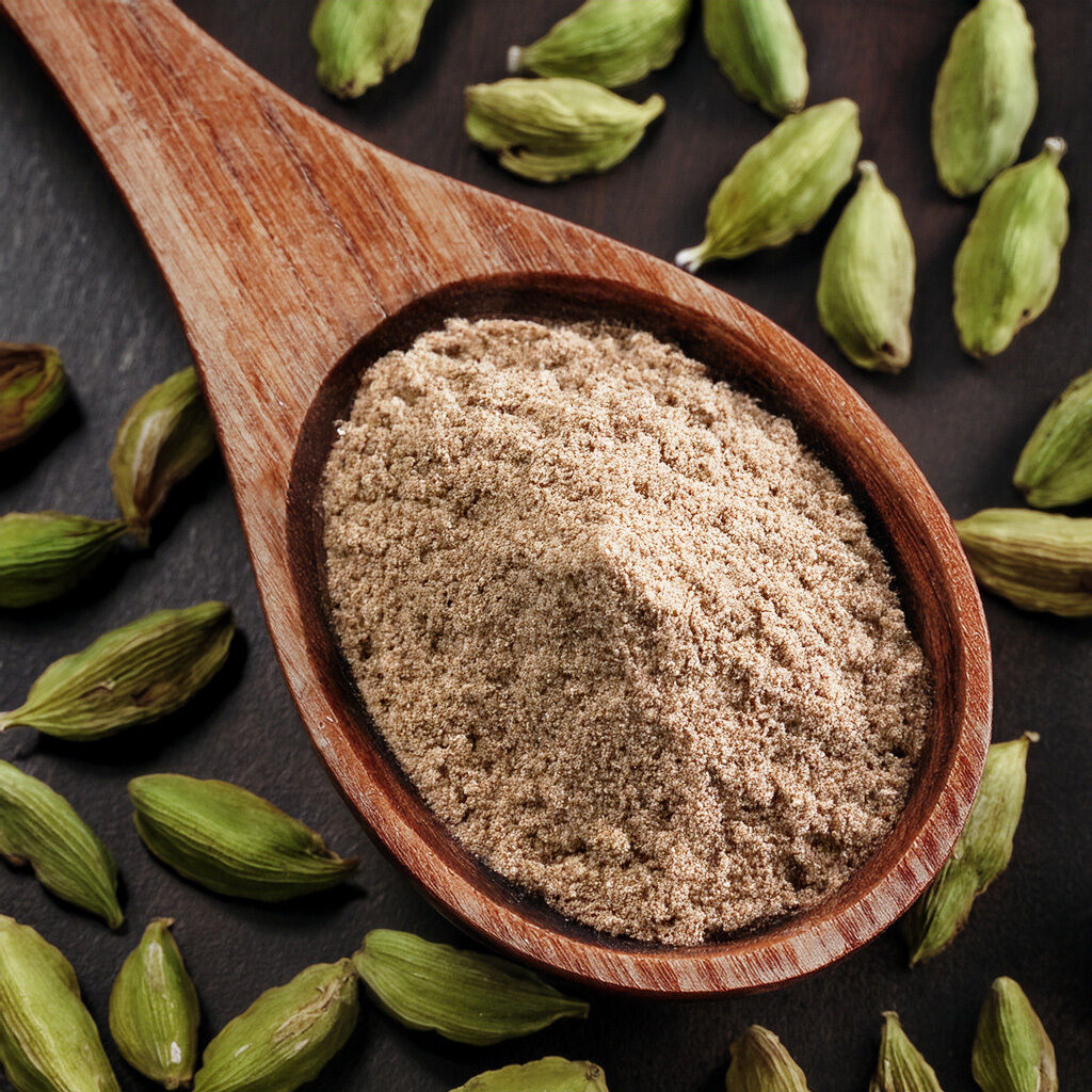 Træske fyldt med lysebrun stødt kardemomme omgivet af grønne kardemommekapsler på mørk baggrund