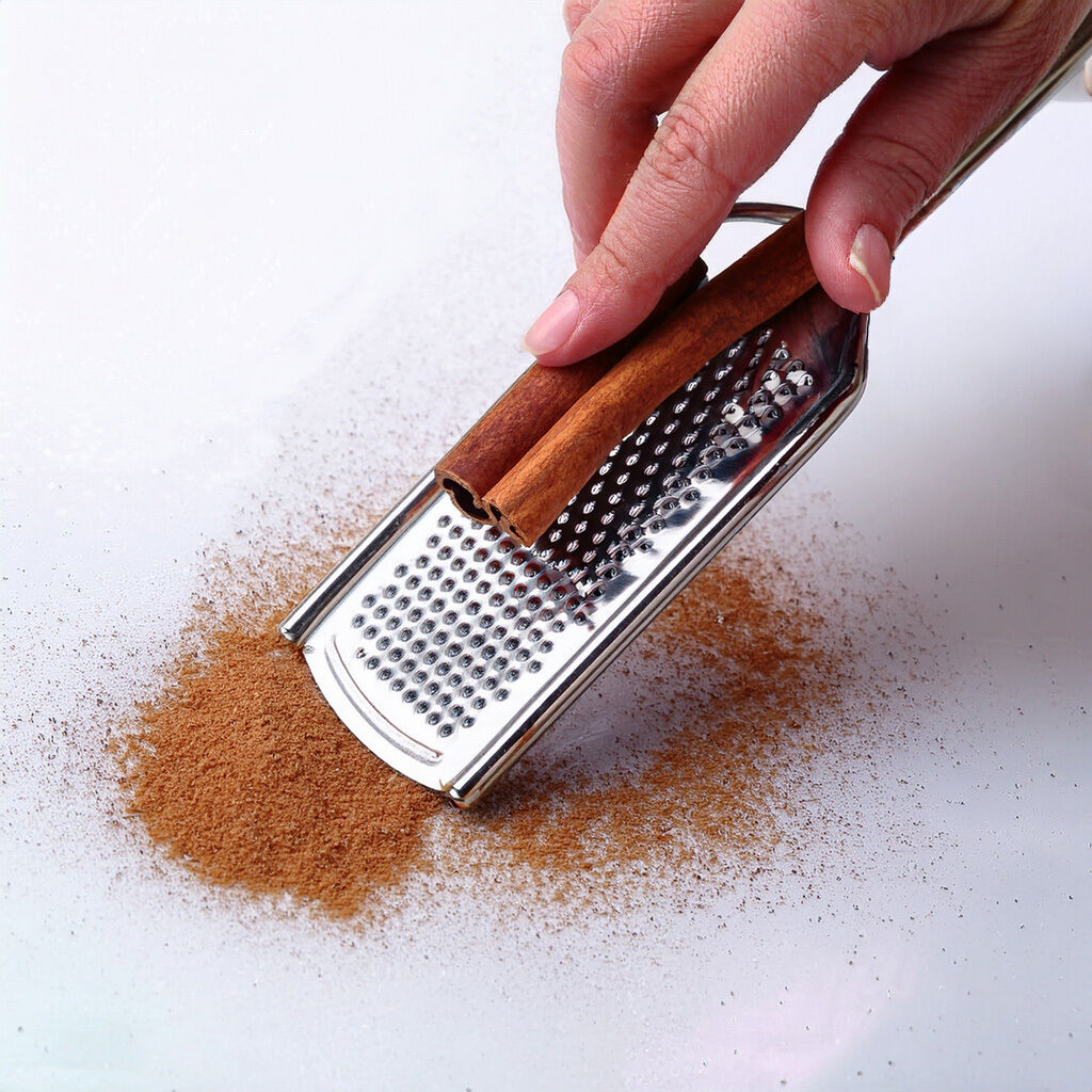 Hånd river en kanelstang over et lille metalrivejern, med friskrevet kanel drysset på en hvid overflade