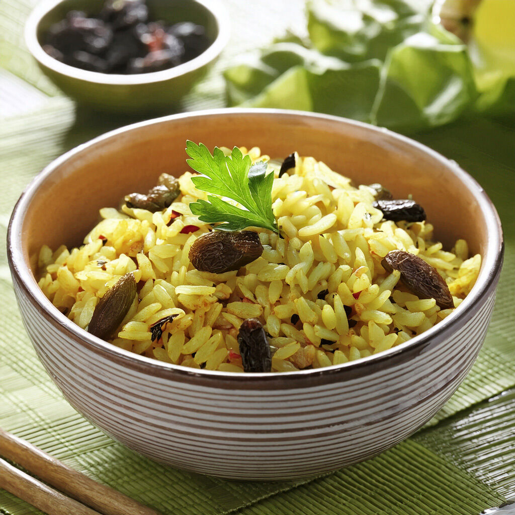 En skål med krydret gul ris, pyntet med persille og rosiner, på en bambusmåtte.