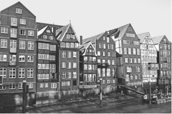 Sort-hvidt foto af en række historiske, smalle pakhuse langs en kaj med refleksioner i vandet