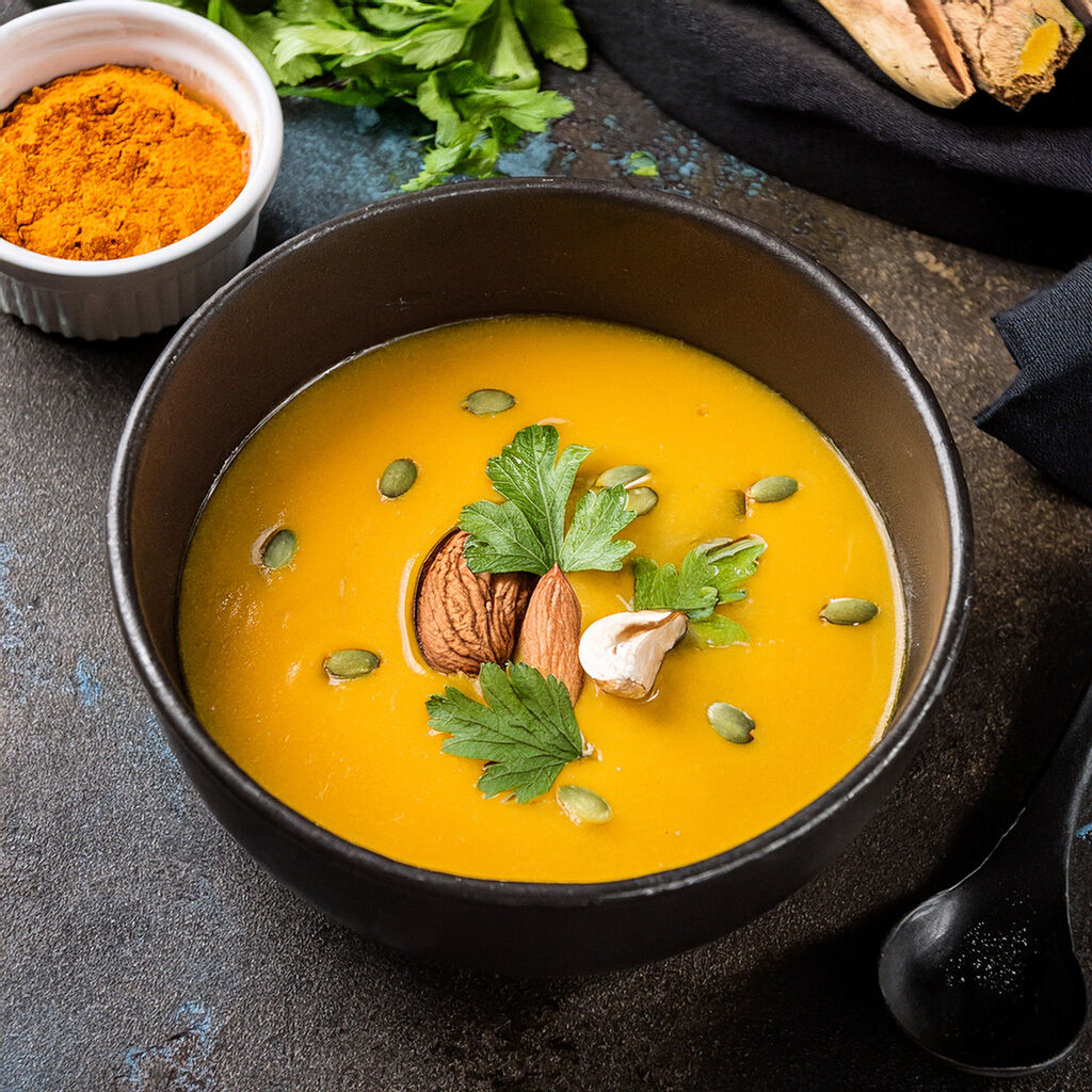 Tæt billede af cremet græskarsuppe i sort skål, pyntet med græskarkerner, hel mandel, et fed hvidløg og friske persilleblade