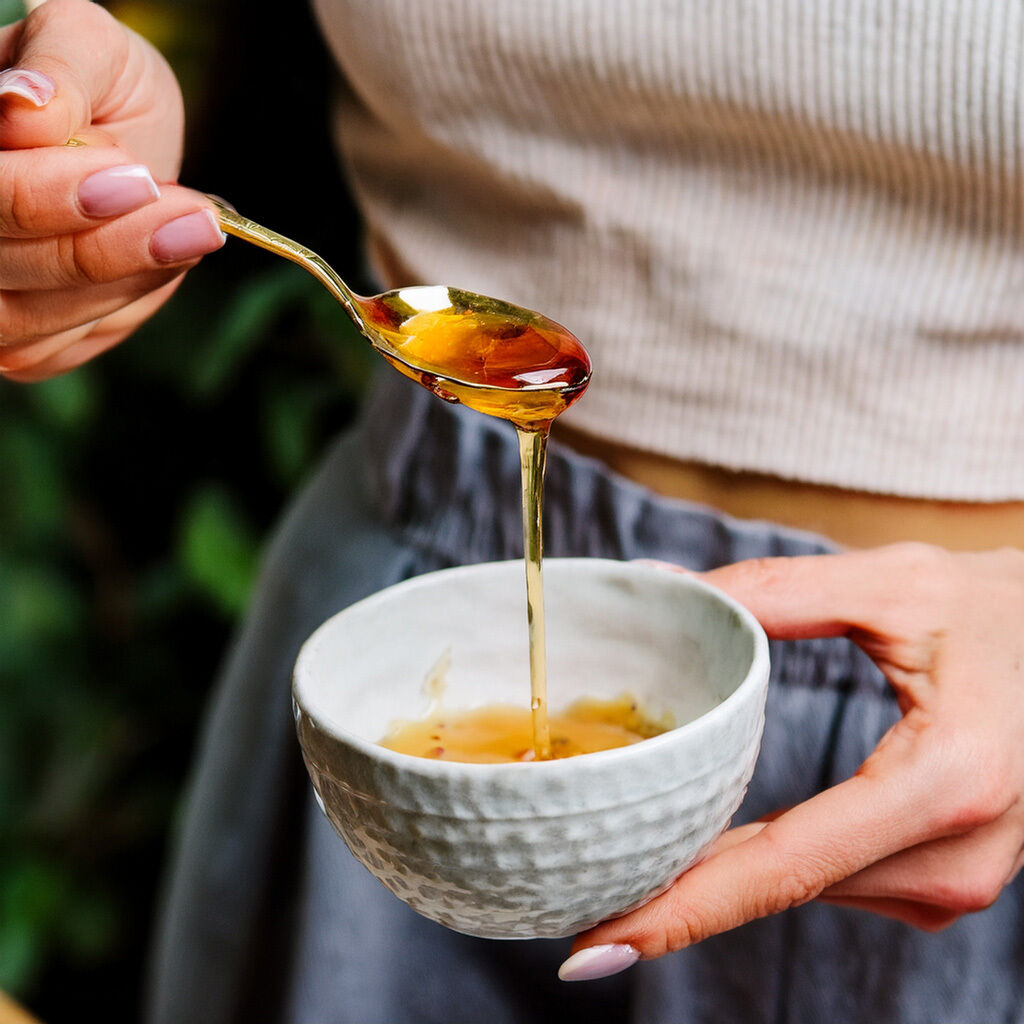 Nærbillede af gylden honning, der drypper fra en ske ned i en hvid keramisk skål, holdt af hænder