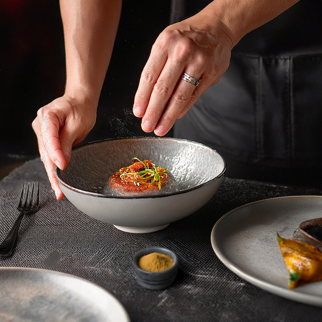 Nærbillede af kokkehænder, der drysser krydderi over en anrettet gourmetret i en keramisk skål på et mørkt bord med bestik og en lille krydderiskål ved siden af.