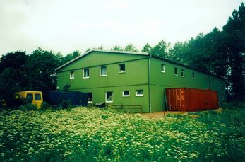 Grøn to-etagers midlertidig kontor- eller lagerbygning i metal på en byggeplads, med orange fragtcontainer og gul varevogn, omgivet af græs og vilde blomster.