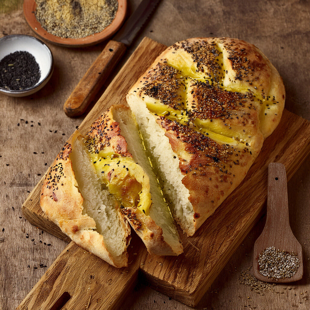 Nærbillede af nybagt, gyldent brød skåret i skiver på et træskærebræt, drysset med sorte og hvide frø, med små skåle og en træske med ekstra frø ved siden af